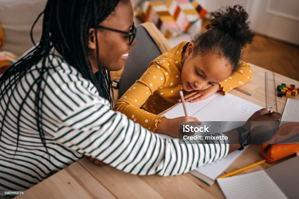 Mother assisting daughter in writing homework at home Mid adult mother with dreadlocks assisting daughter in writing homework on table at home Homework Stock Photo