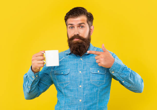 Serious man pointing finger at coffee mug yellow background, copy space.