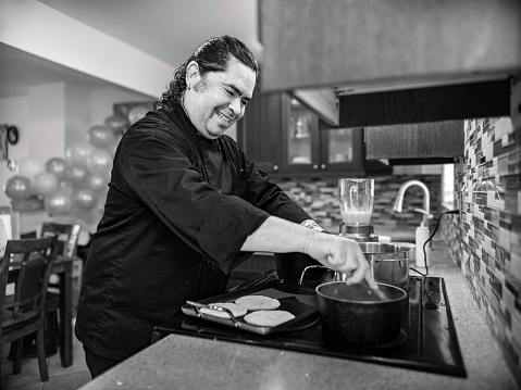 LGBTQ Mexican Male Chef in his kitchen cooking a meal. He is dressed in black chefs robe. Interior of kitchen in private city home.