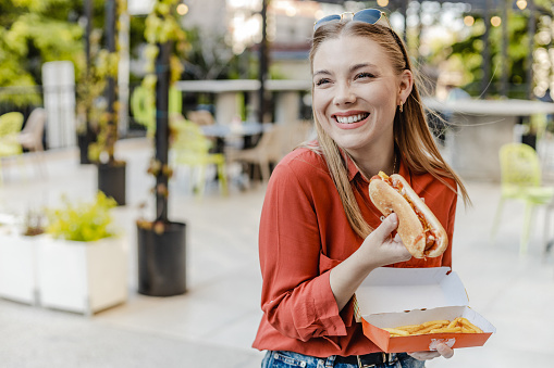 A Caucasian young woman is eating a hot dog and smiling