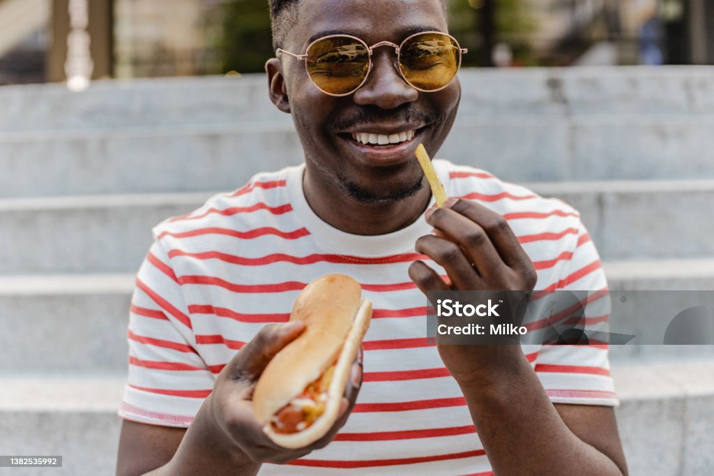 Portrait of a young African-American man is eating hot dog and smiling A young modern man is eating a hot dog and smiling Eating Stock Photo