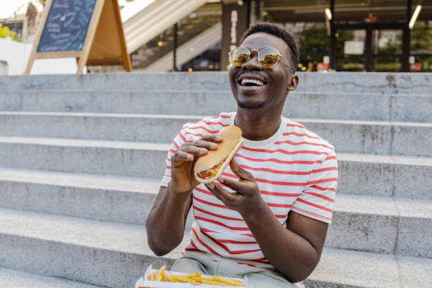 Young African-American man is eating hot dog and smiling stock photo