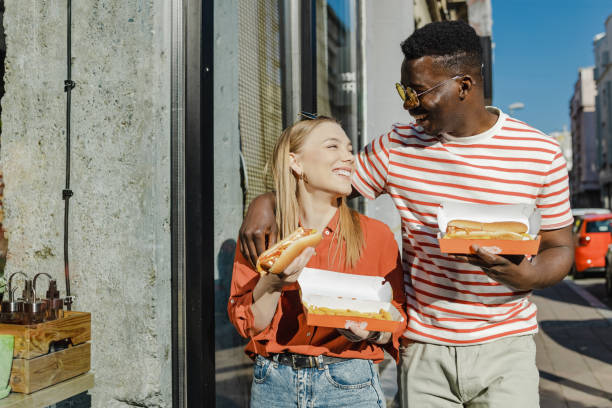 i giovani mangiano hot dog e camminano per strada - couple blond hair social gathering women foto e immagini stock