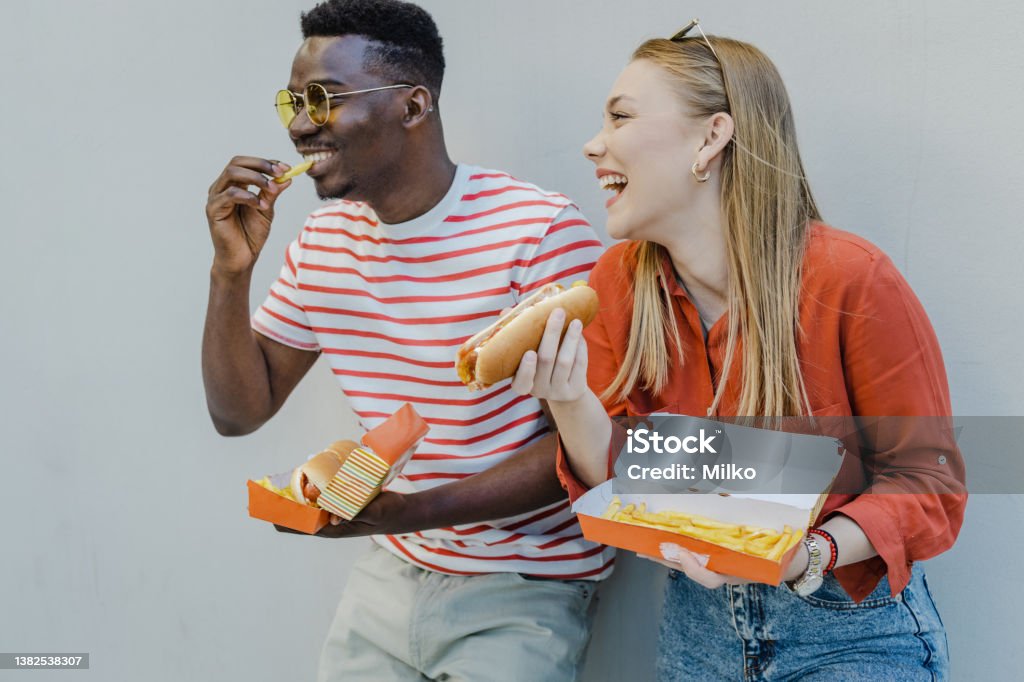 Hanging out and eating hot dog with a friend outdoors African American man and a caucasian young woman are eating a hot dog in front of the wall Hot Dog Stock Photo
