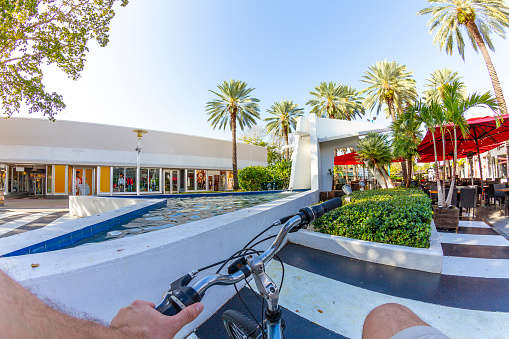 POV Point of view shot of a young latin sportsman biking a bicycle at Lincoln Road Mall, a touristic open shopping mall center street, part of Art Deco District near to Ocean Drive, South Beach, Miami Beach, Miami, South Florida, United States of America - USA. \n\nShooting from a personal perspective POV in an exotic tropical beach travel holidays after Corona Virus Pandemic Illness Covid-19 finished.