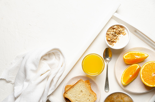A cup of coffee, orange juice, some toasts, pieces of orange, and a bowl of plain yoghurt with granola in a white tray with a white napkin, on a white background.