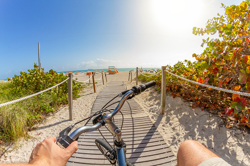 POV Point of view shot of a young latin sportsman tourist biking a bicycle to the beach at South Beach, Miami Beach, Miami, South Florida, United States of America - USA after Corona Virus Pandemic Illness finish.

On background, the famous and touristic Art Deco District. 

Shooting from a personal perspective in an exotic tropical beach travel holidays.
