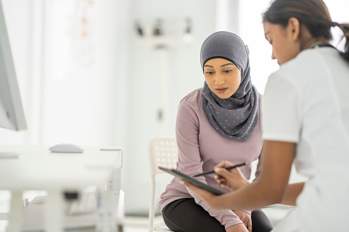 A female doctor sits with a Muslim patient as she reviews her recent test results on a tablet with her.    She is seated in white scrubs as she explains the results on the tablet screen to the patient.  The patient is seated on a chair in front of her and leaning in to look closely.