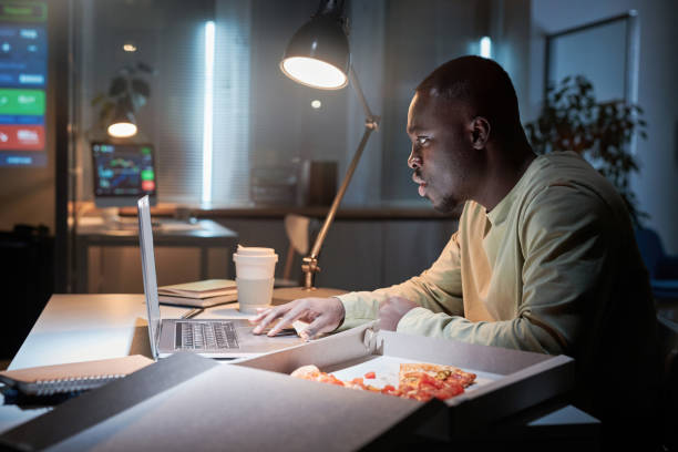 Man working at office during deadline African businessman sitting at workplace and having a snack with pizza and coffee while working on laptop at office till late night working overtime stock pictures, royalty-free photos & images