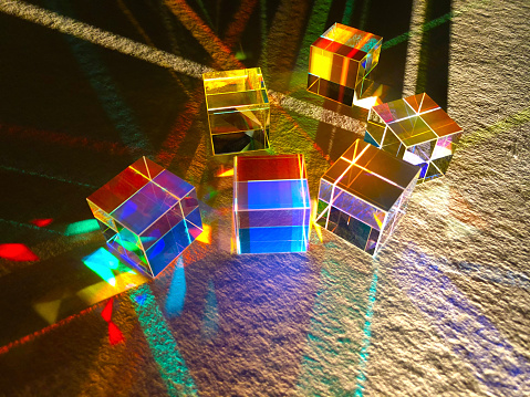 Narrow beam of artificial light shines through stack of dichroic coloured glass cubes and spreading into several spectrum colours. Reflection and refraction of light and lighting. Colourful stripe pattern on texture white paper.