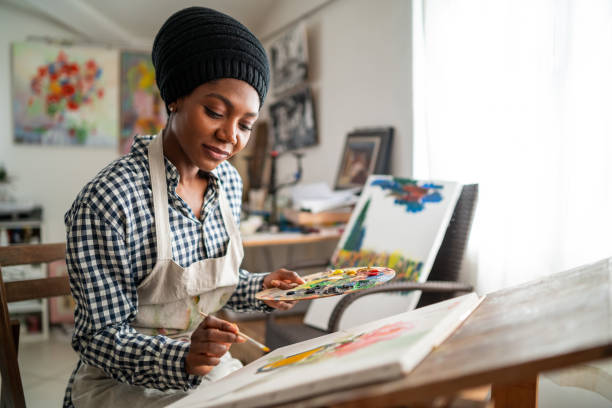 Afro female fine artist with traditional african hat 
 and apron, drawing in art studio stock photo