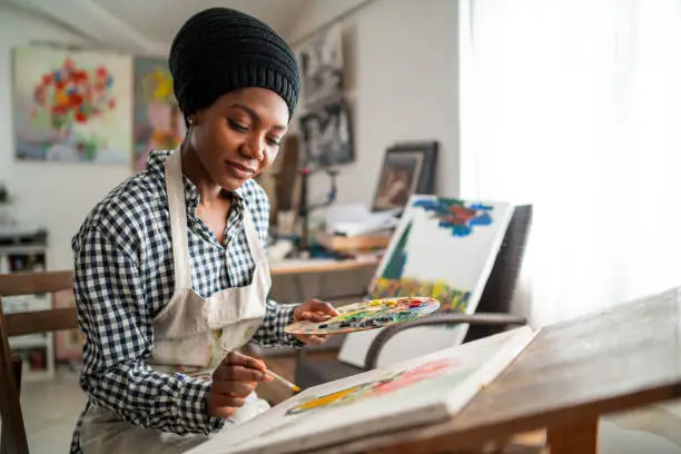 Photo of Afro female fine artist with traditional african hat 
 and apron, drawing in art studio