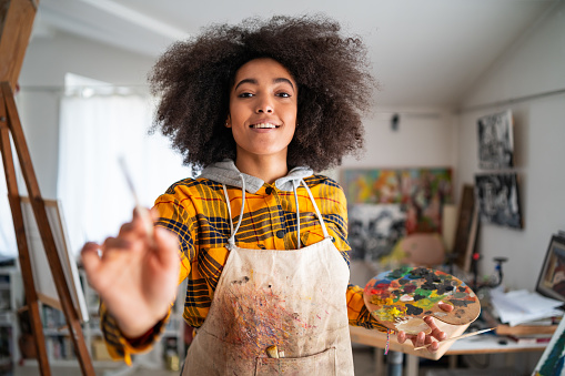 Portrait of smiling afro female fine artist with afro hair, wearing apron and holding paintbrush, standing in an art studio