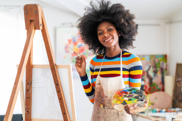 sonriente artista femenina africana con cabello afro y con un delantal, dibujando en un estudio sobre un lienzo, colocado en un trípode - entertainment school art studio painter fotografías e imágenes de stock