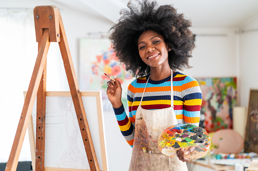 Smiling african female fine artist with afro hair and wearing an apron, drawing in a studio on a canvas, placed on a tripod