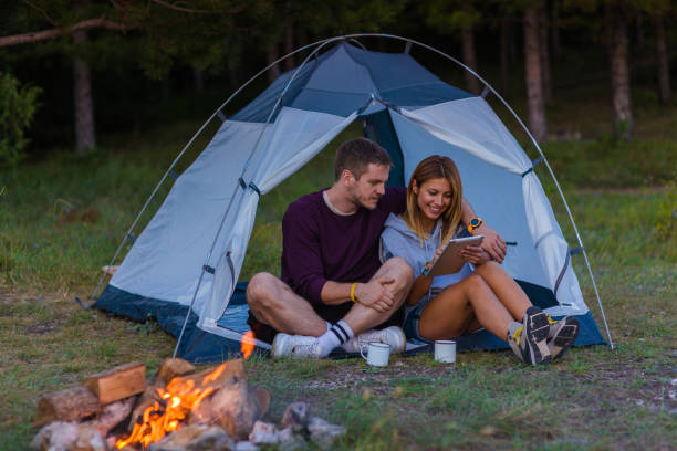 istock Young couple drinking coffee, enjoying the mountain view and checking the tablet with bonfire in the sunset 1382884798