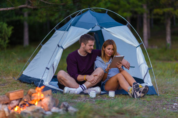 istock Young couple drinking coffee, enjoying the mountain view and checking the tablet with bonfire in the sunset 1382884800