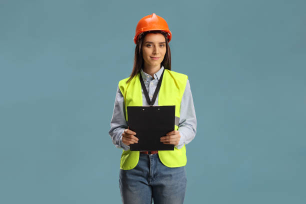 Young female engineer with a safety vest and hardhat holding a clipboard Young female engineer with a safety vest and hardhat holding a clipboard isolated on blue background waistcoat stock pictures, royalty-free photos & images