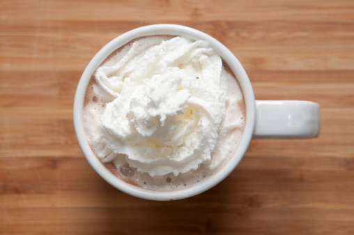 A cup of cocoa with whipped cream, sitting on a wooden surface, looking down from above.