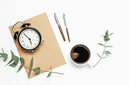 Flat lay cup of black coffee, black alarm clock, notebook, diary, eucalyptus on white background top view copy space. Minimalistic food concept, Feminine desk workspace, time to work coffee background.
