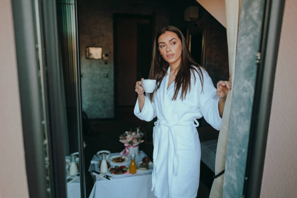 mujer tomando café y mirando a través de la ventana de la habitación del hotel - bathrobe women cup towel fotografías e imágenes de stock