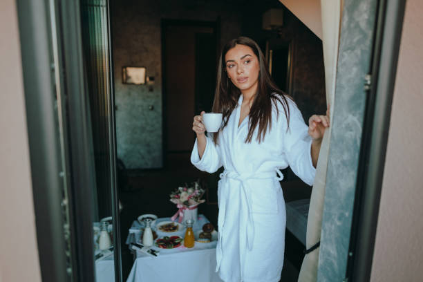 donna che beve caffè e guarda attraverso la finestra della camera d'albergo - bathrobe women cup towel foto e immagini stock