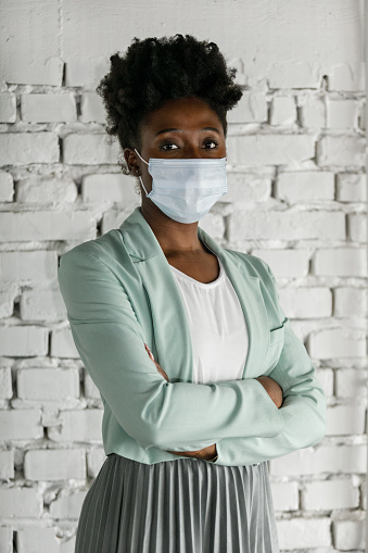 Portrait of confident, young Black businesswoman standing with arms crossed, against white brick wall, wearing a protective face mask and looking at camera.