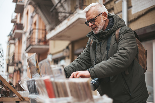 Senior man chooses vintage vinyl records at a flea market. Buys rare audio recordings, nostalgia.