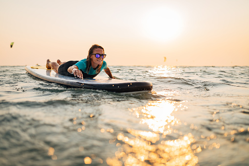 Lying on a SUP board, a cheerful woman surfing the sea. She loves playing with the waves, the open sea and the excitement that this extreme sport brings.