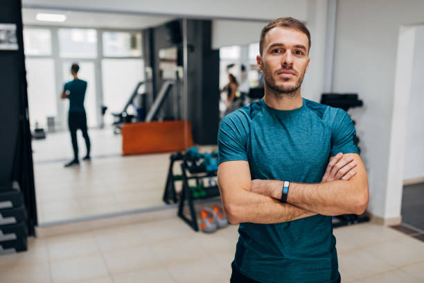 ein hüfthoher blick auf einen athletischen, motivierten mann, der mit verschränkten armen in der turnhalle steht. - waistup stock-fotos und bilder