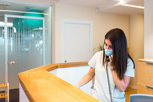 Female receptionist with a face mask attending at a call while working at the front desk of a health center. Health care concept.