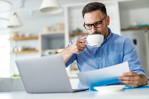 Young smiling businessman drinking coffee while working on laptop and documents at home.