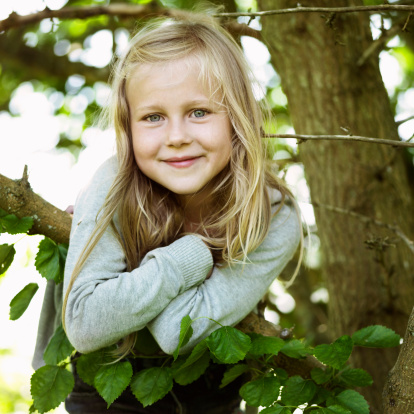 A child is playing with some daisies in the forest. She is tired, so she rests on the ground leaning against an old tree.