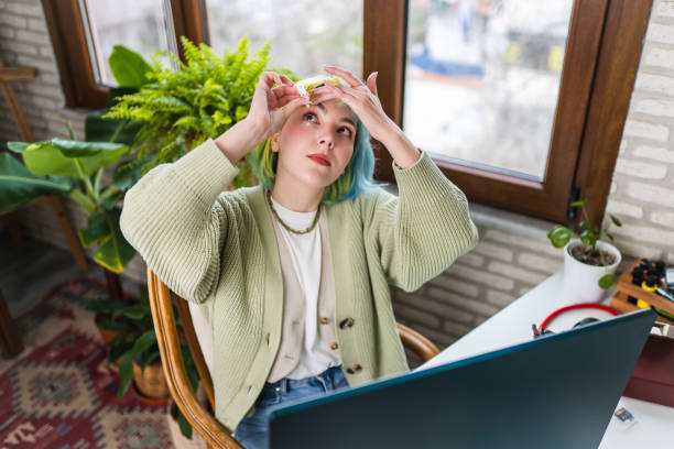 Young woman with dry eye uses eye drops while working stock photo