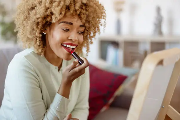 Happy curly african american woman putting lipstick on at home