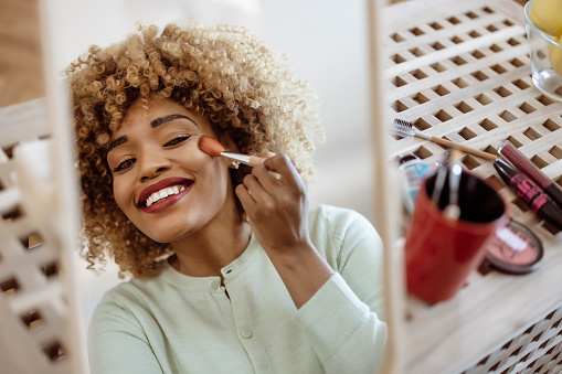 Reflection in the mirror of happy curly woman makes face contouring, holds makeup brush