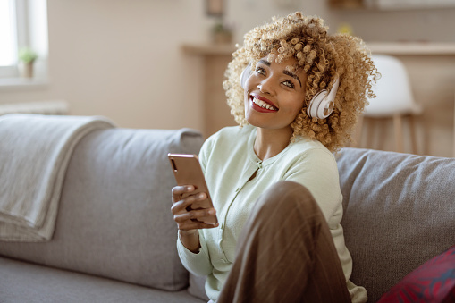 Beautiful afro american woman in headphones is listening to music, using a smart phone and smiling while sitting on couch at home