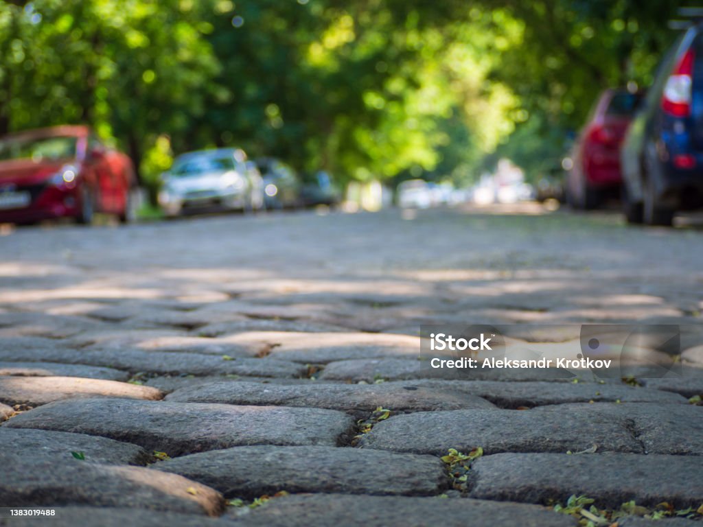 A city alley paved with cobblestones and parked cars in the shade of trees in a quiet, peaceful area. Focus on the foreground. A European paved street lined with cars A city alley paved with cobblestones and parked cars in the shade of trees in a quiet, peaceful area. Focus on the foreground. A European paved street lined with cars. City Street Stock Photo