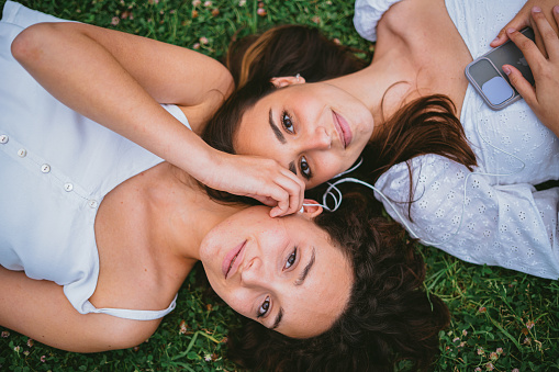 Two teenager girls looking at camera while they are listening to music. One of them is holding the earphone. They are relaxing on grass.