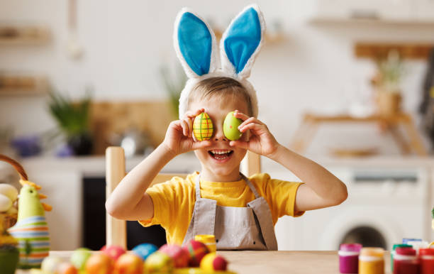 istock Funny happy kid boy with plush bunny ears on head covering eyes with multicolored Easter eggs 1383028055