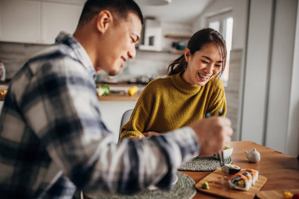 Couple eating sushi at home Two people, modern Japanese couple having sushi together for lunch in dining room at home. mid adult couple stock pictures, royalty-free photos & images