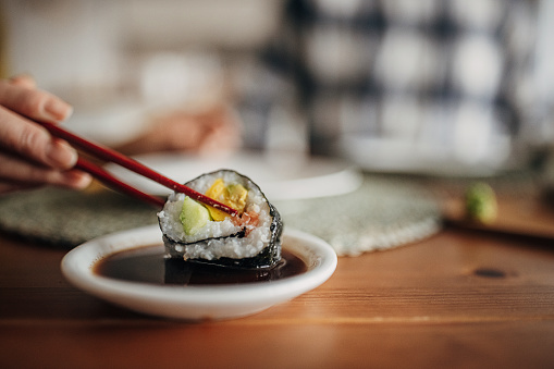 Two people, modern Japanese couple having homemade sushi together for lunch in dining room at home. Woman is dipping sushi in soy sauce.