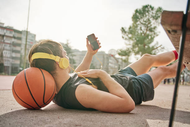 istock basketball player lying down after training. leaning on basketball. making a video call with smart phone with yellow earphones. 1383035112