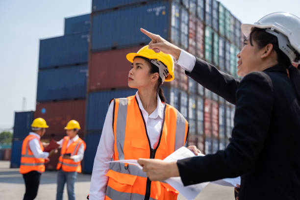 istock woman worker working with Foreman, standing with ware a yellow helmet to control loading and check a quality of containers from Cargo freight ship for import and export at shipyard or harbor 1383053477