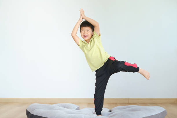 Cute funny Asian kindergarten boy child try to practices yoga in Tree Pose and meditating at home stock photo