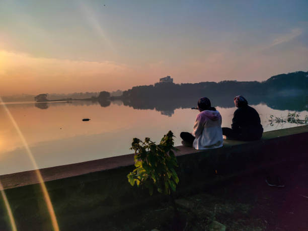 Stock photo two young Indian girl sitting near lake and enjoying beautiful sunrise in the winter season, Picture captured at Rankala lake , Kolhapur, India. Kolhapur, India- December 5th 2020; Stock photo two young Indian girl sitting near lake and enjoying beautiful sunrise in the winter season, Picture captured at Rankala lake , Kolhapur, India. kolhapur stock pictures, royalty-free photos & images