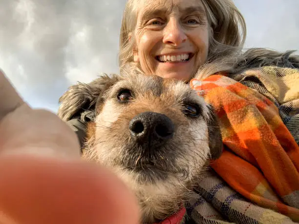 A selfie shot of a mature woman wearing warm clothing with her senior patterdale terrier while standing outdoors in Northumberland. They are both looking at the camera and the woman is smiling.