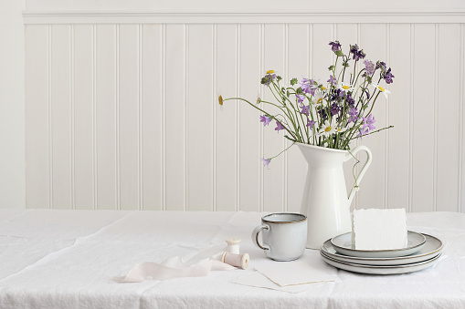 Spring, summer breakfast still life. Jug with wild flowers bouquet on linen table cloth. Daisies, bluebells, aquilegia. Greeting card mockup, cup of coffee, ceramic plates. White wooden wall backround.