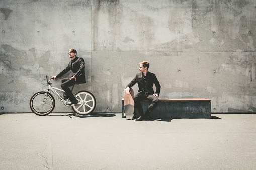 twin brothers in business suits sitting on a bench with skateboard and on a bmx cruiser bicycle in urban surrounding.