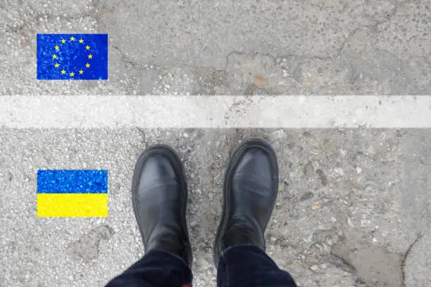 A person in winter boots standing on asphalt next to Ukraine flag, border and EU flag. Politics, refugees, war crisis and humanitarian disaster concept.